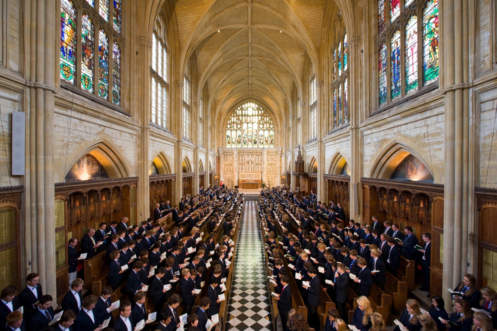 A large group of people in formal attire stand in rows inside a grand, ornate chapel at Cheltenham College Boarding School, with high arched ceilings and colourful stained glass windows, holding papers and facing the altar at the front.