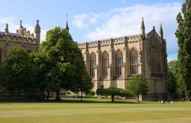 A large, historic stone building—part of Cheltenham College Boarding School—with arched windows and spires rises behind green lawns and tall trees; a few people are sitting on the grass under a partly cloudy sky.
