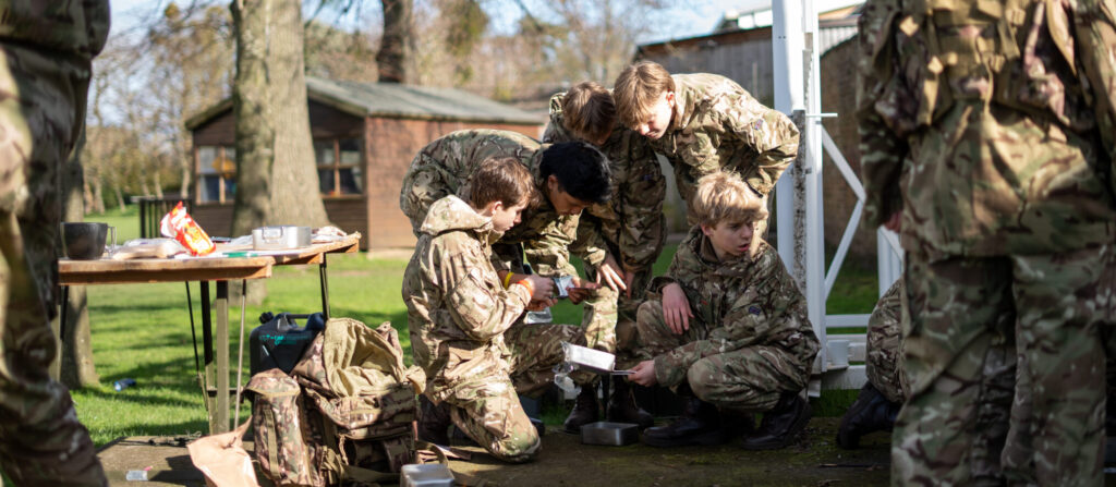 A group of young people in camouflage uniforms, part of the Combined Cadet Force, crouch together outdoors examining equipment on the ground. Other members stand nearby, with a wooden building and trees visible in the background on a sunny day.