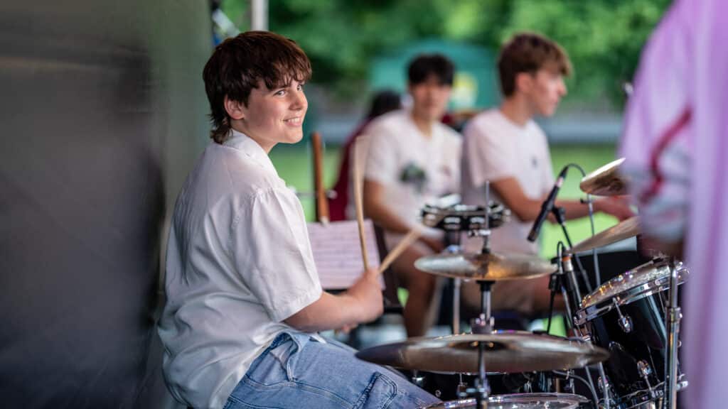 A smiling boy in a white shirt and ripped jeans plays drums on an outdoor stage at CCFM 2025: College Community Music Festival, whilst two other boys watch, surrounded by greenery and sunshine.