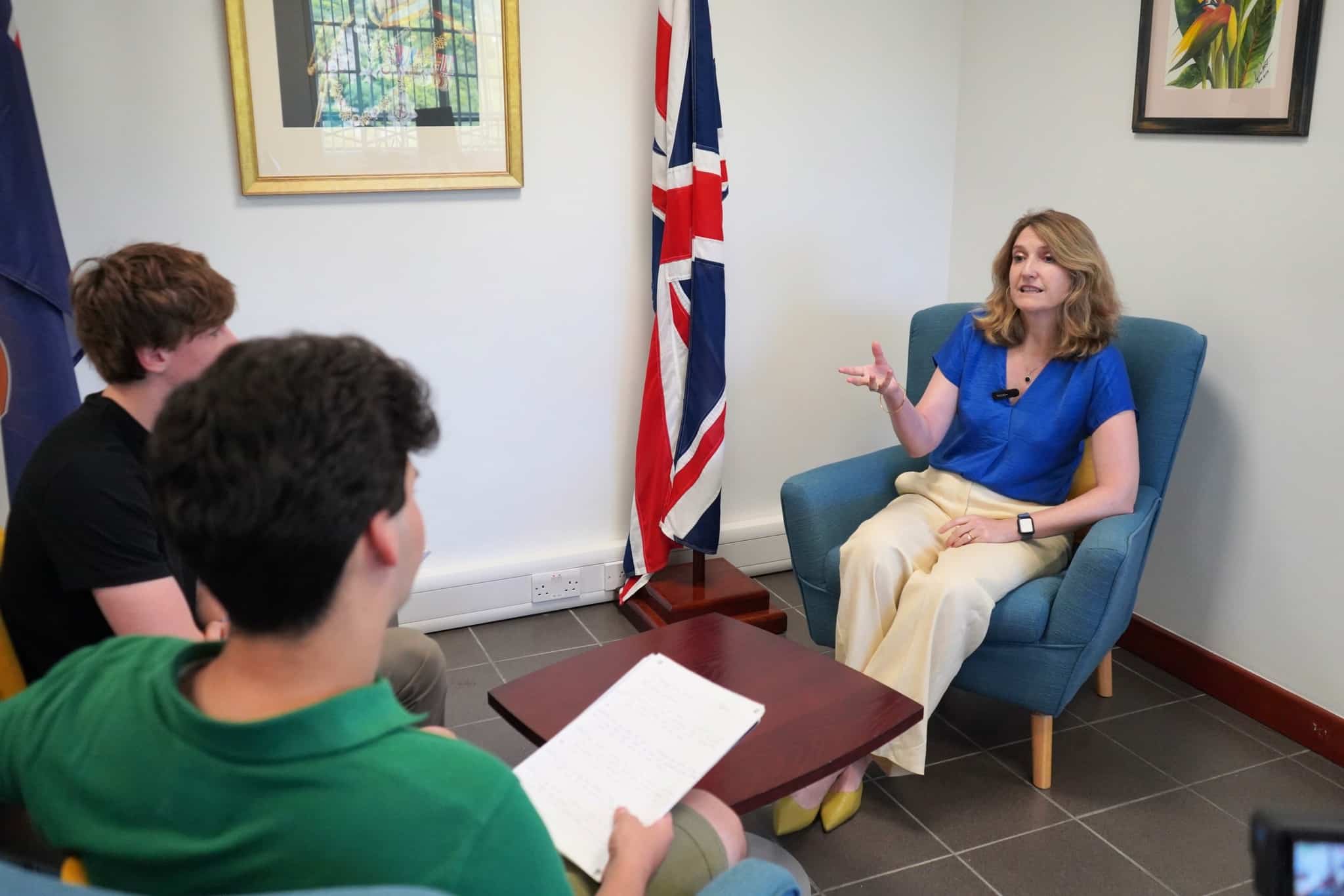 A woman sits in a blue armchair speaking to two young men, one holding a notepad. A British flag is displayed behind her in a brightly lit office, as they discuss learning from leadership in Montserrat amid framed pictures on the walls.