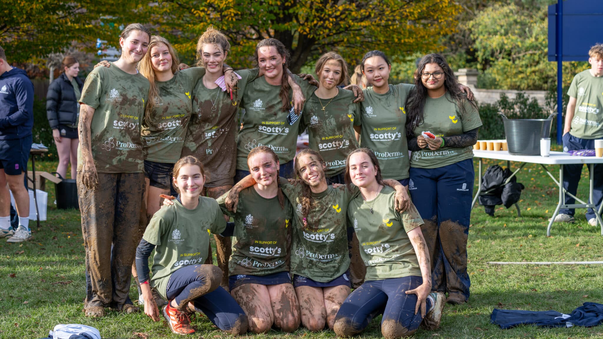 A group of eleven young women in muddy sports kit smile and pose together outdoors on a grassy pitch, suggesting they have just finished the Cheltenham College Charity Mud Run. Trees with autumn foliage and people are visible in the background.