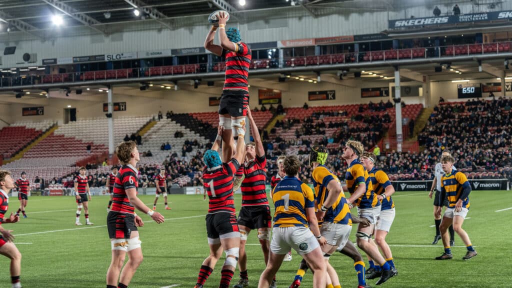 Rugby players in red and black, and yellow and blue jerseys compete in a line-out during the Cheltenham College Charity Rugby Match 2025, as one player is lifted to catch the ball in a stadium with spectators in the background.