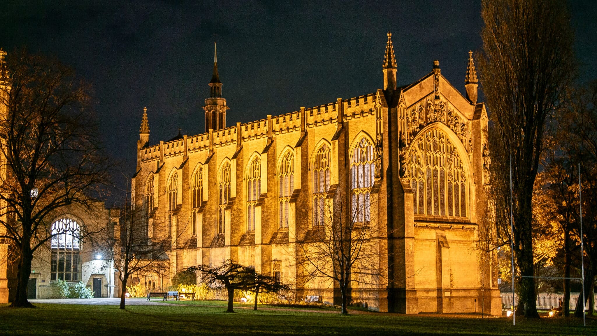 A large, historic, Gothic-style building—home to Cheltenham College Higher Education & Careers—illuminated by golden lights at night, with arched windows and spires, surrounded by trees and a grassy lawn.