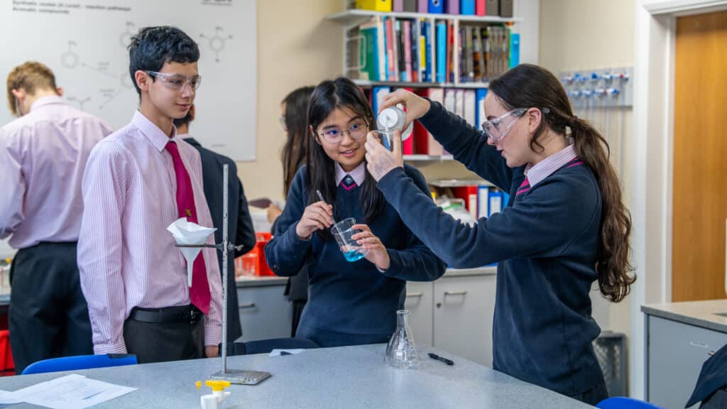 Three Lower College students in school uniforms and safety goggles conduct a science experiment in a classroom, pouring liquid into a beaker, with lab equipment and books visible in the background.