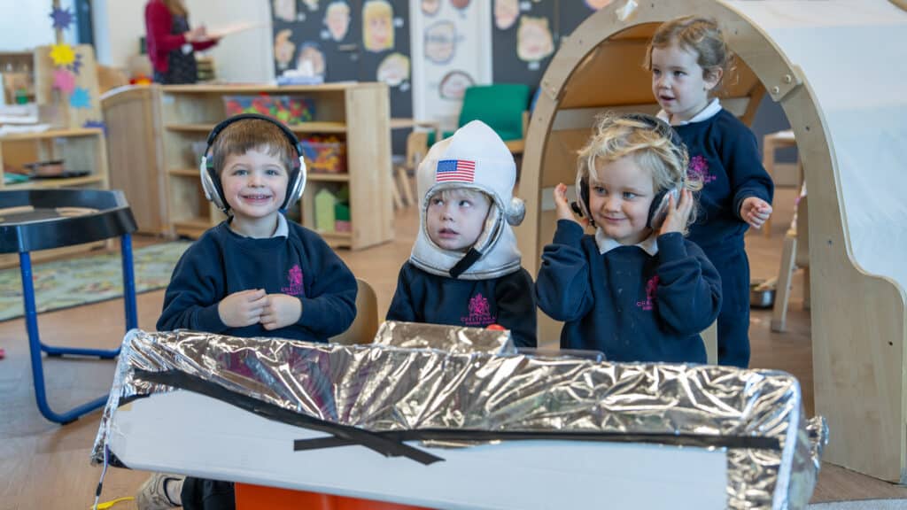 Four young children at Cheltenham Nursery School pretend to be astronauts. Three wear headphones, while one sports a space helmet with an American flag. They sit behind a cardboard and foil spaceship, smiling and playing together.