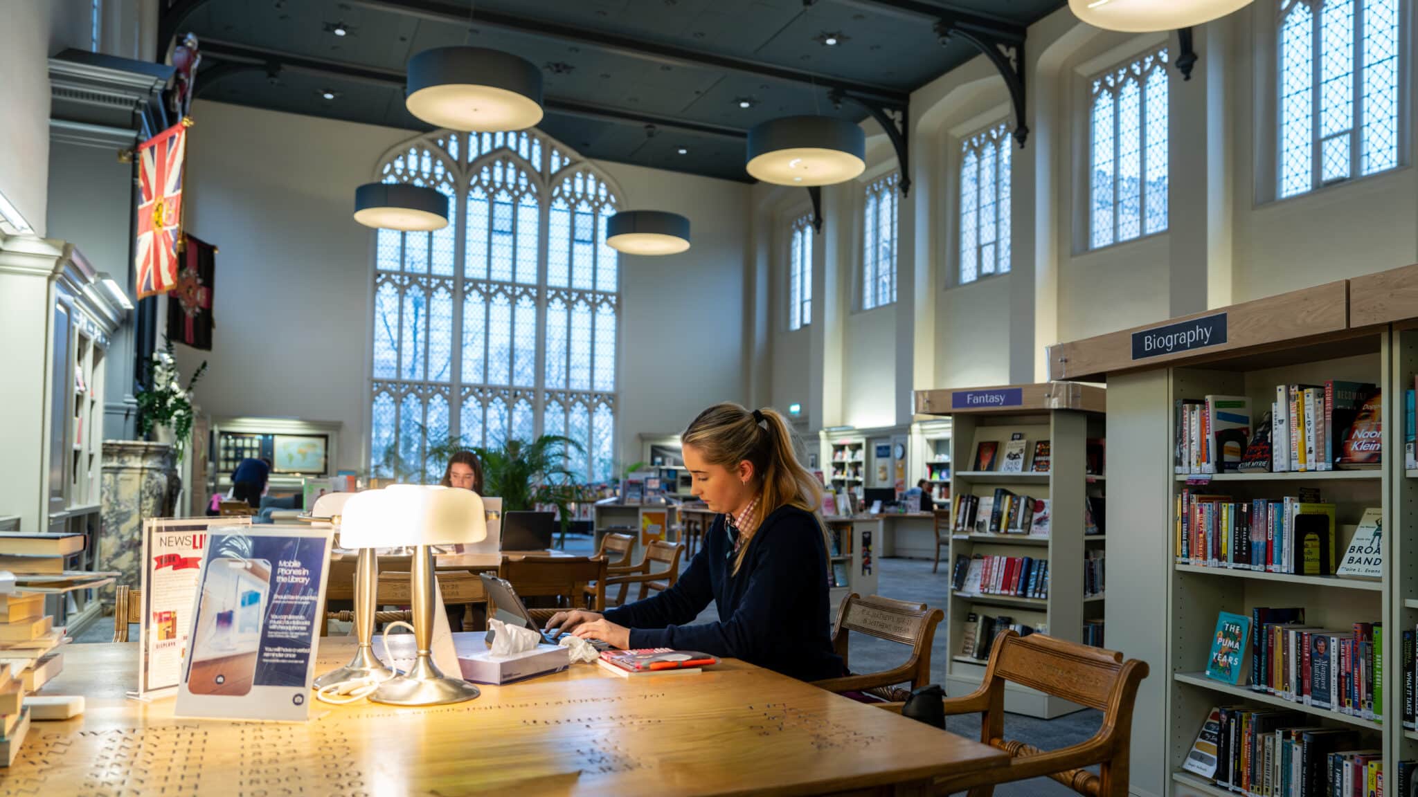 A woman sits at a wooden table in a spacious library with large arched windows, reading and writing about Sixth Form Subjects. Bookcases, lamps, and people studying are visible in the background, creating a quiet academic atmosphere.