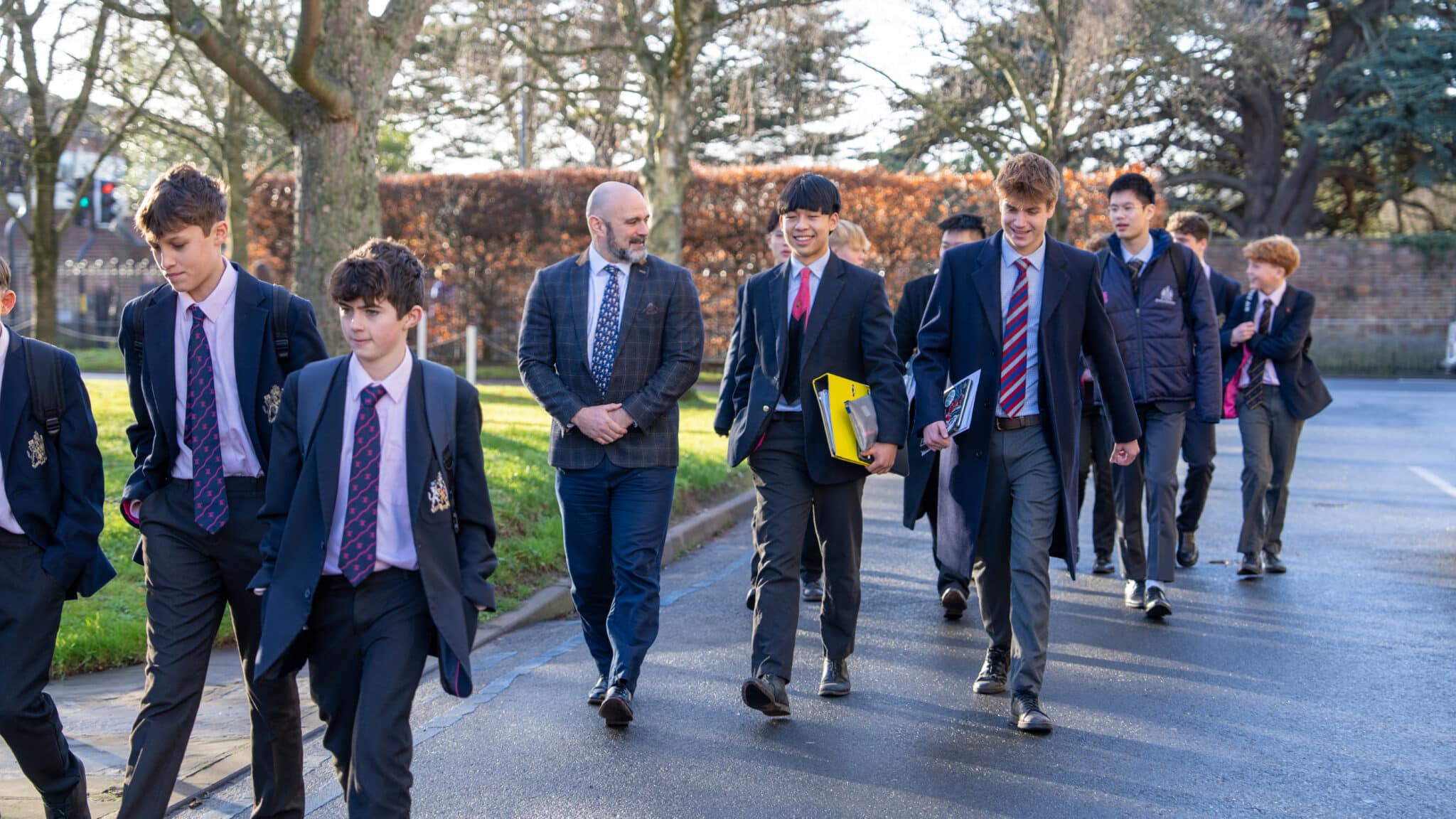A group of schoolboys in uniforms walk outside with a male teacher on a sunny day, carrying folders and chatting about their Sixth Form subjects. Trees and a brick wall are in the background.