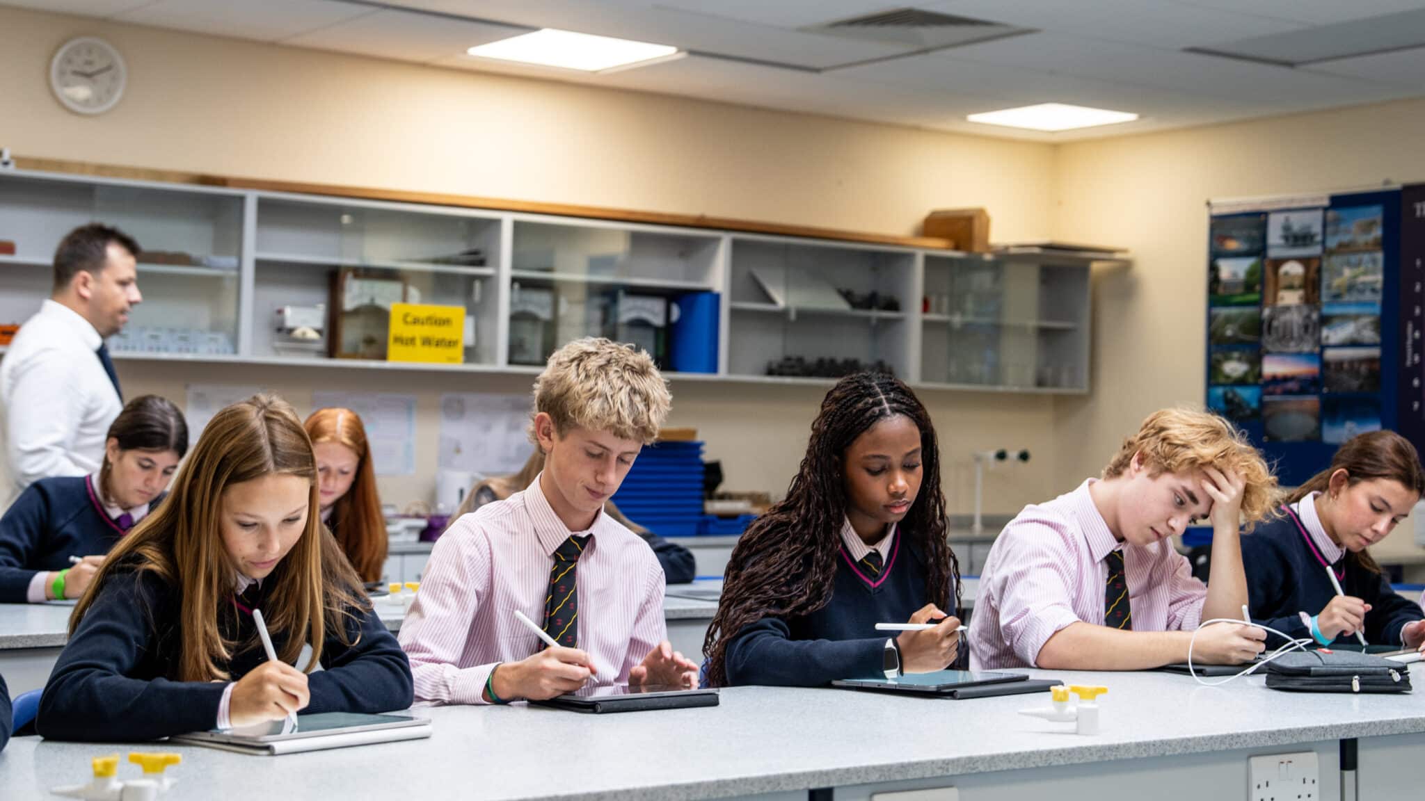 A group of students in school uniforms sit at desks using tablets in a classroom, exploring Sixth Form subjects, whilst a teacher stands in the background. The classroom features shelves, lab equipment, and educational posters.