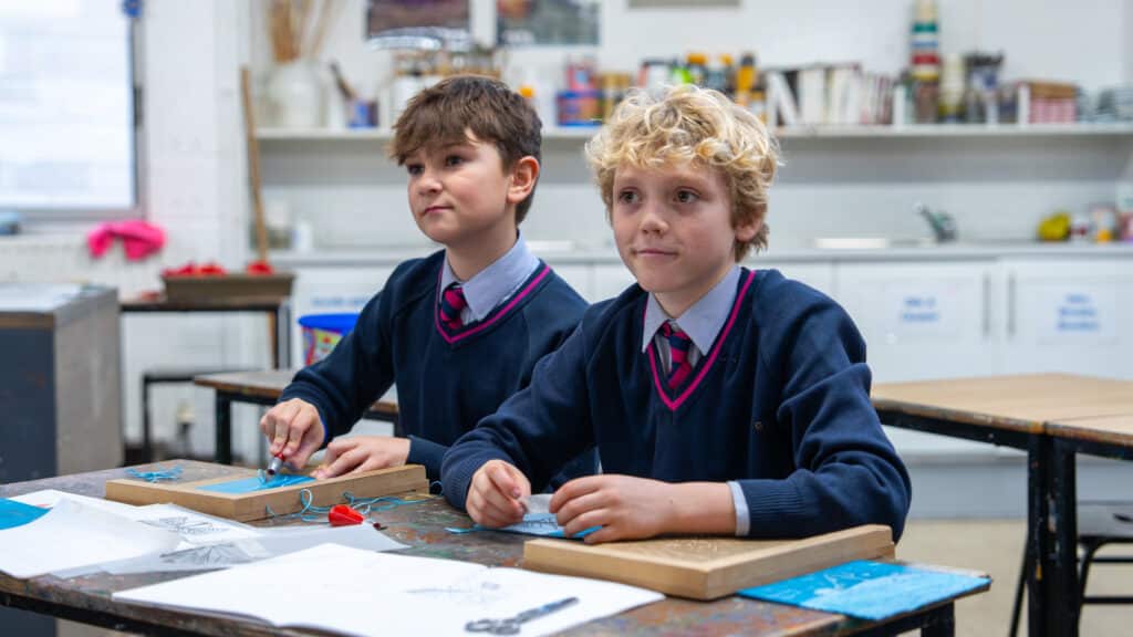 Two boys in school uniforms sit at a classroom desk, engaged in a co-curricular project with wooden boards and wires. Papers, tools, and art supplies are scattered around, reflecting their hands-on learning experience.