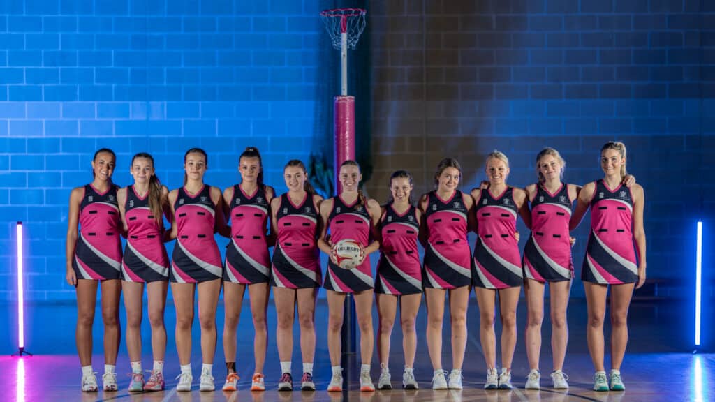 The Cheltenham College Charity Netball 2026 team of 10 women in matching pink and black kits stands in a row indoors, smiling at the camera, with a netball post and blue lighting behind them. One player in the centre holds a netball.