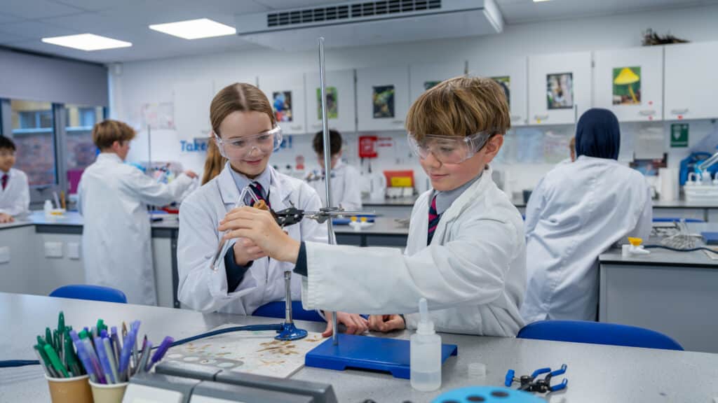Two Prep Year 3 - Year 8 pupils in lab coats and safety goggles work together on a science experiment, while classmates and a teacher stay busy in the background. Laboratory equipment and supplies are visible on the tables.