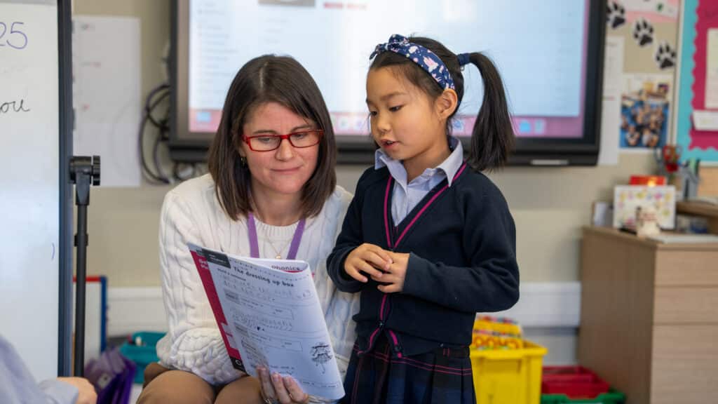 A teacher wearing glasses sits next to a young girl in a classroom, holding and discussing a worksheet. The girl stands, listening closely—perfect for Reception to Year 2 learning—with classroom supplies and a screen in the background.