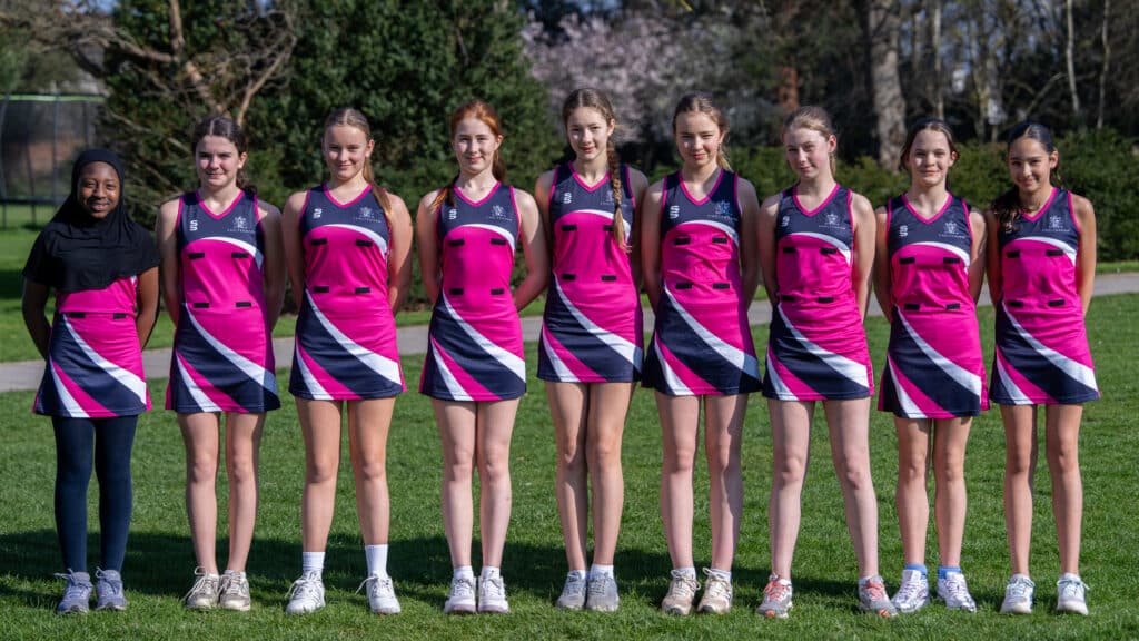 Nine girls in matching pink, black, and white uniforms stand in a line on the grass, smiling at the camera during the Cheltenham Prep Charity Netball Match 2026, with trees and sunshine in the background.