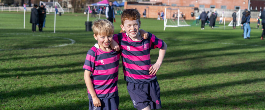 Two young boys in matching pink and navy striped football kits stand arm in arm on a grassy pitch at the Cheltenham Prep Football Festival, smiling at the camera. Other children and adults are visible playing and standing in the background.