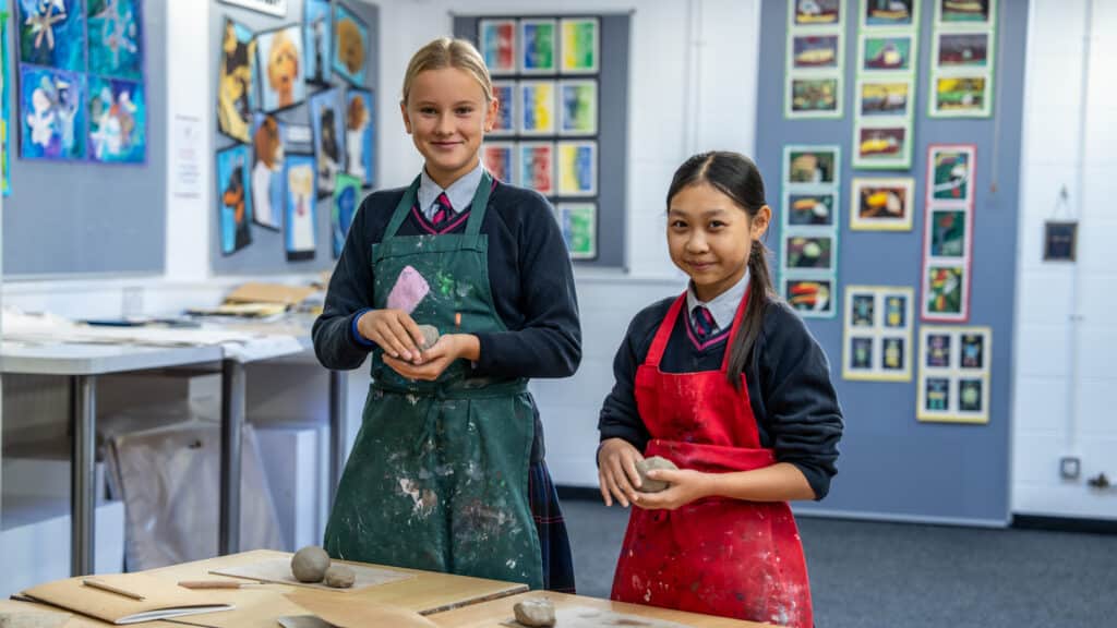 Two schoolgirls in aprons stand at a table in an art classroom at Cheltenham Day School, smiling and working with clay. Colourful artwork is displayed on the walls behind them.