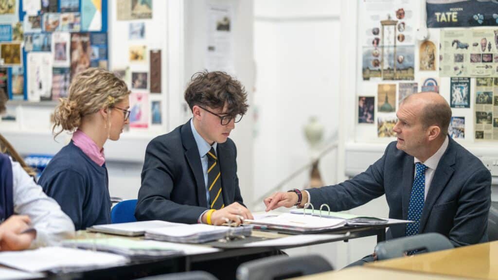 A teacher in a suit sits at a desk with two students, discussing Oxbridge Preparation papers in a classroom decorated with various posters and images on the walls.