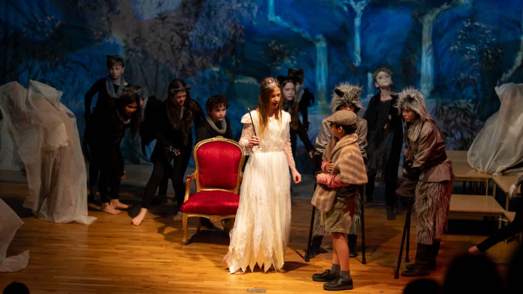 A group of children in costumes perform on stage during a Cheltenham Prep Drama Performance; a girl in a white dress and crown stands by a red chair, surrounded by kids dressed as Narnia animals, with a boy in a cap and brown coat before her.