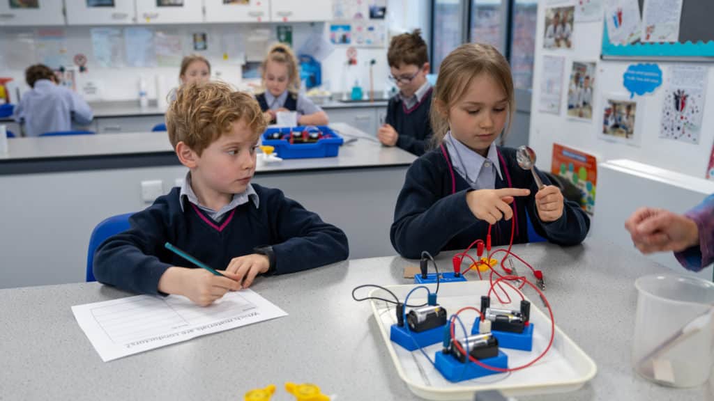 A group of young pupils in Cheltenham Prep uniforms work on a British Science Week 2026 experiment with batteries, wires, and bulbs at a classroom table. One girl connects wires while a boy writes on a worksheet, as others work in the background.