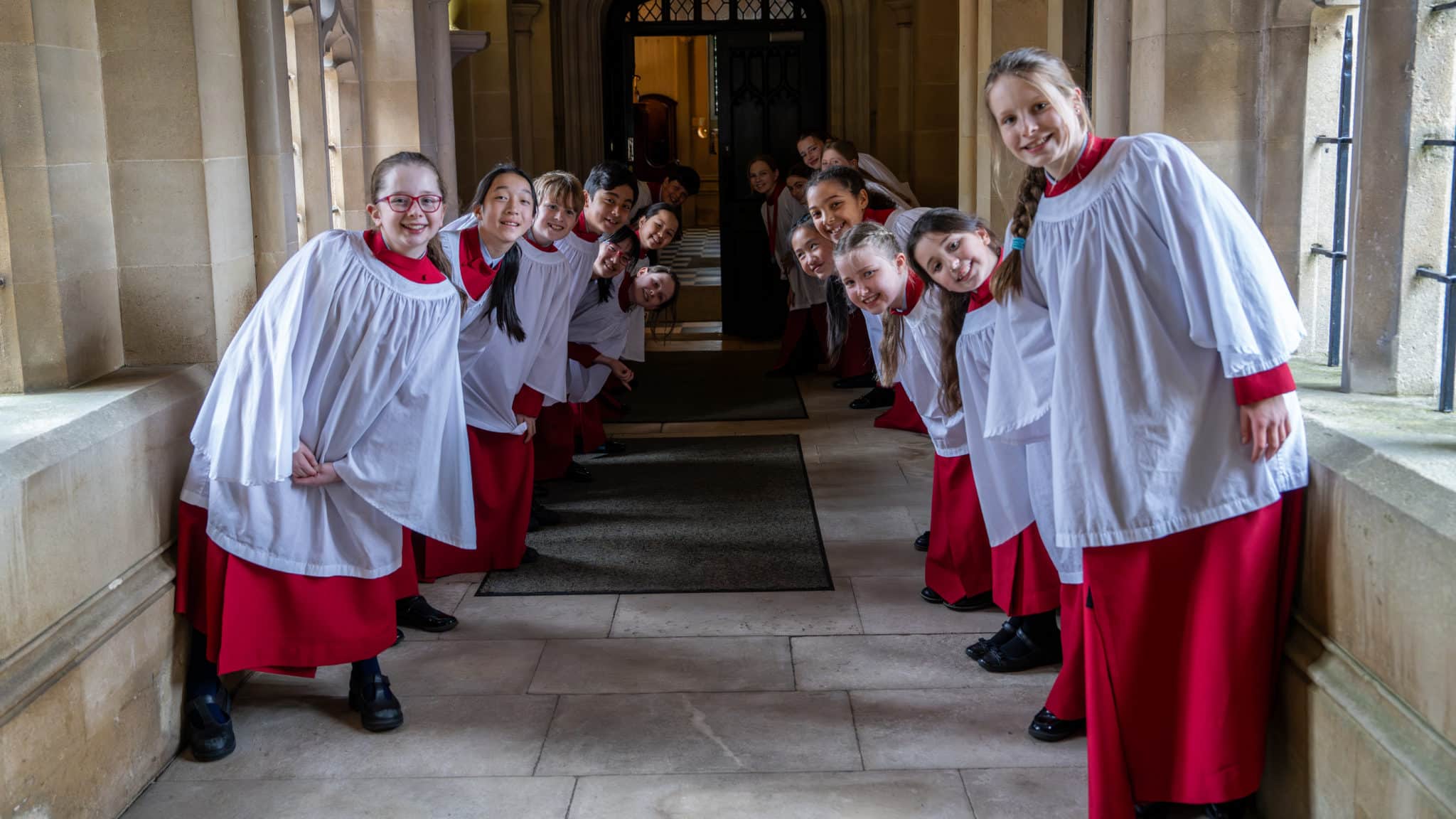 A group of choir children in red robes and white surplices stand in two lines facing each other, smiling and leaning inwards, inside a stone corridor with arched windows—one of Cheltenham Prep Spring Term Highlights 2026.