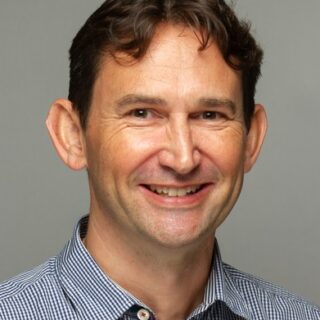 A man with short brown hair and a light complexion is smiling at the camera. He is wearing a checked button-up shirt and is posed against a plain grey background, reflecting the welcoming spirit of Cheltenham College Boarding School.