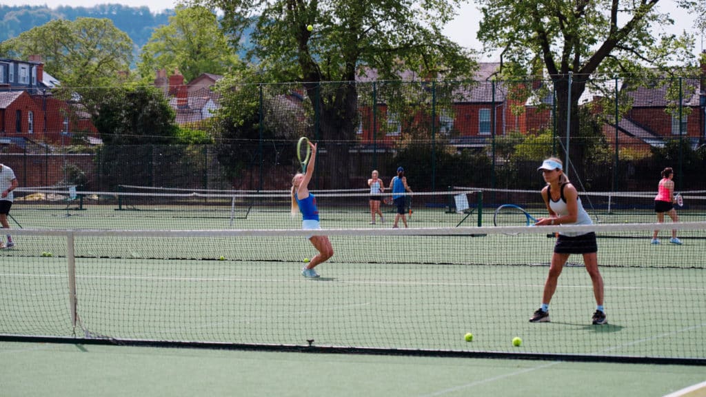Two women play tennis on an outdoor court; one is preparing to hit the ball, while the other is ready near the net. Other people are visible playing on courts in the background, with trees and houses behind them.