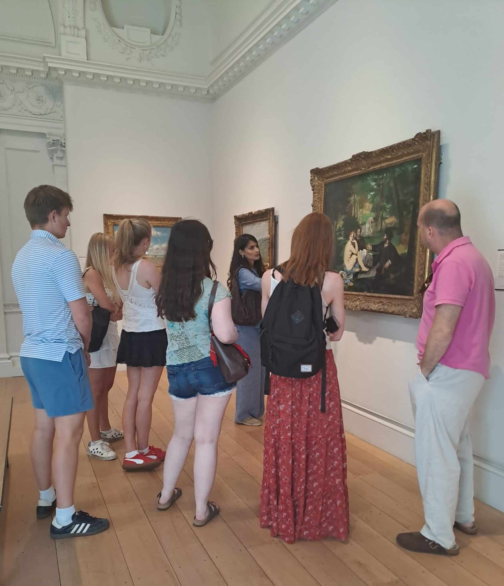 A group of people stands in an art gallery during History of Art Futures Week, closely observing framed paintings on a white wall. One person appears to be explaining or discussing the artwork to the others amid ornate details and wooden floors.