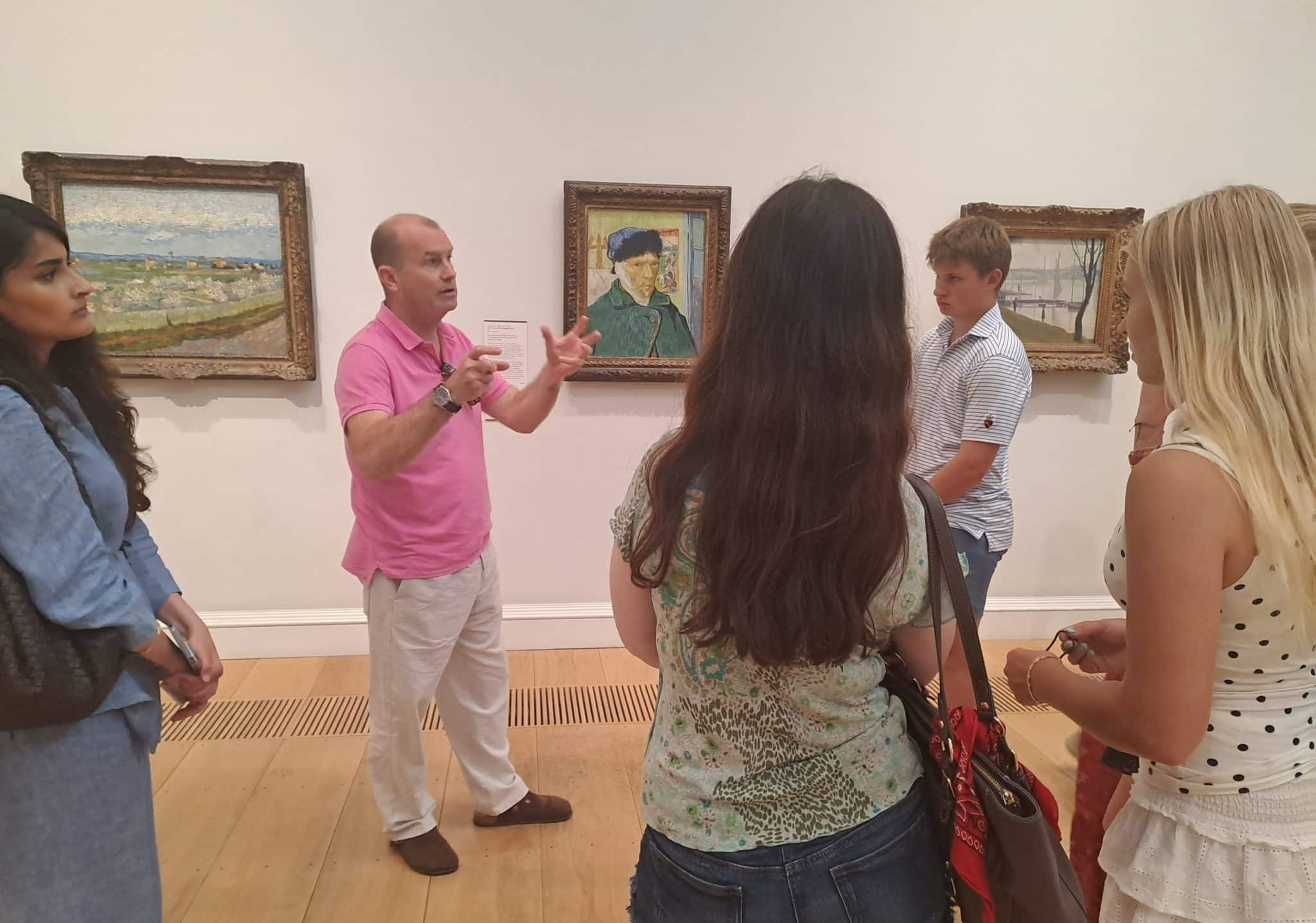 A man in a pink shirt gestures whilst speaking to a group of four people in an art gallery during History of Art Futures Week, with several framed paintings hanging on the wall behind them.