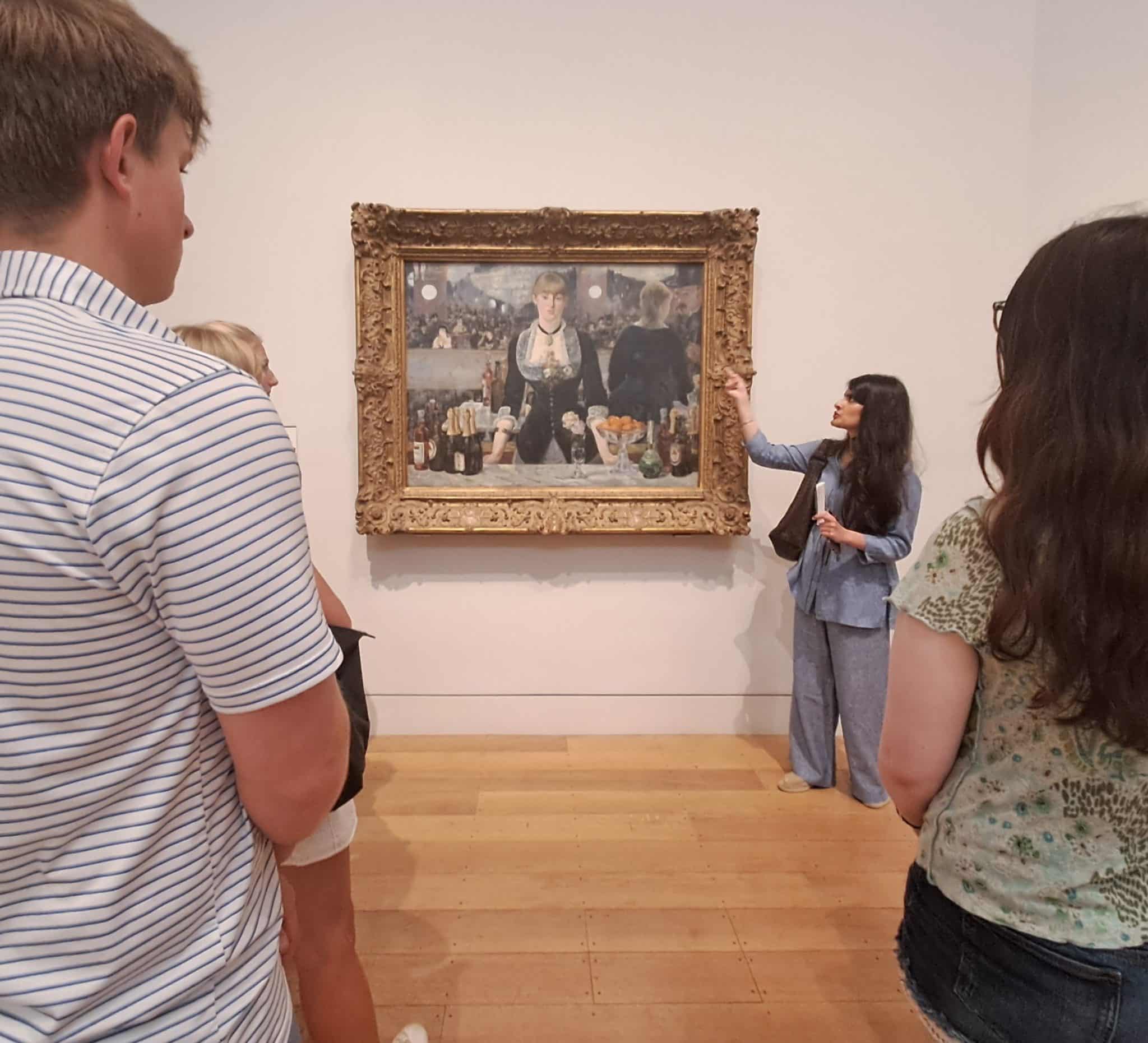 A group of people listens to a woman giving a tour during History of Art Futures Week in an art gallery, standing in front of a framed painting on a white wall. The painting depicts a bar scene with a woman at the centre.