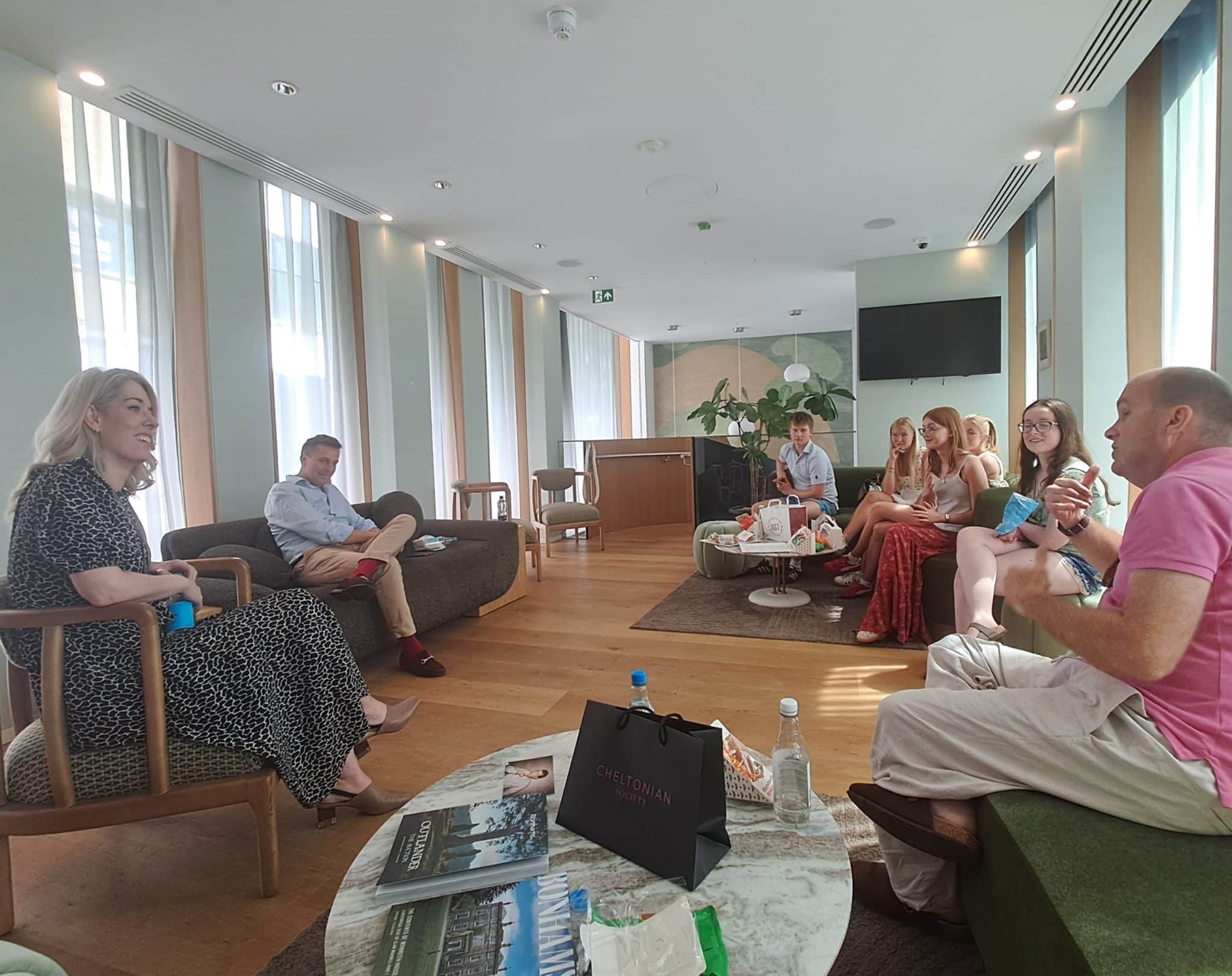 A group of seven people sit and talk in a modern, sunlit lounge with large windows during History of Art Futures Week. Some hold drinks, and bags and magazines are on the table in the foreground. The atmosphere appears relaxed and informal.