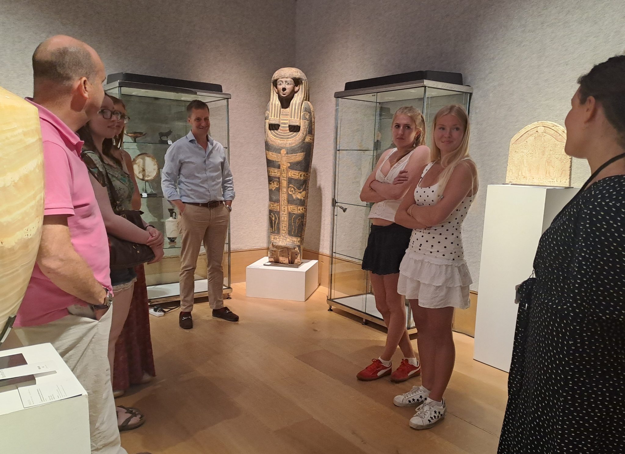 A group of people stands in a museum room around an ancient Egyptian sarcophagus, listening to a guide as part of History of Art Futures Week. The scene is set with wooden floors, soft lighting, and two glass cases displaying artefacts.