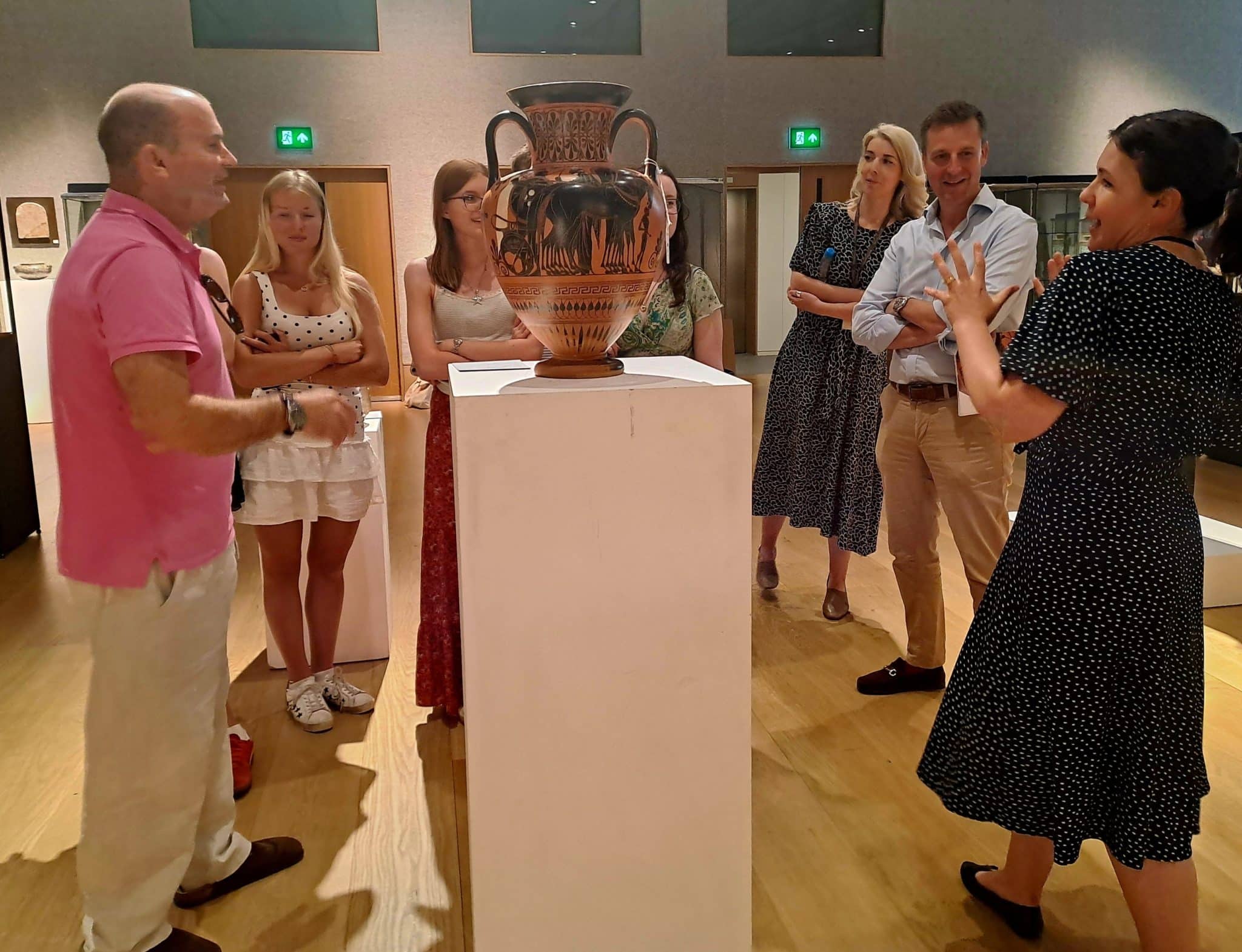 A group of people stands around a large ancient vase displayed on a plinth in a museum, attentively listening to a woman in a black dress as part of History of Art Futures Week, who appears to be explaining something about the artefact.