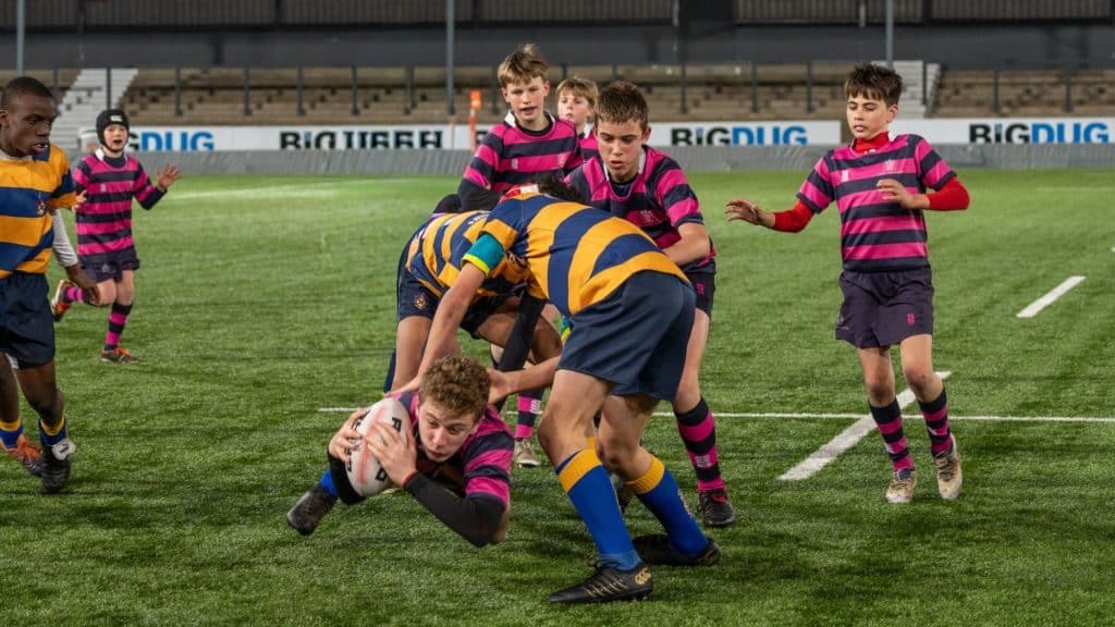 A youth rugby player in a striped jersey dives forward with the ball whilst being tackled by an opponent; other players in Cheltenham College yellow-blue and pink-black jerseys watch on a grassy pitch.