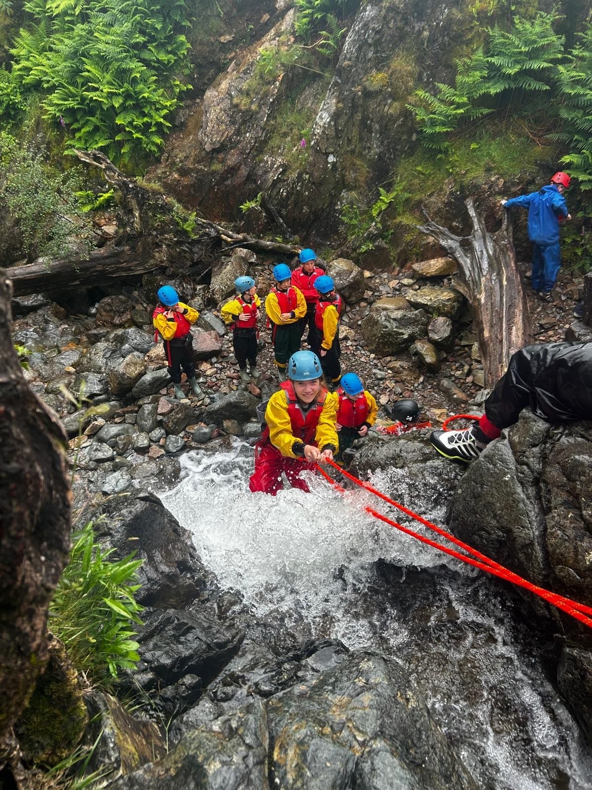 A group of people wearing helmets and waterproof jackets climb a rocky waterfall using ropes, whilst others wait and watch on the rocks below, surrounded by greenery.