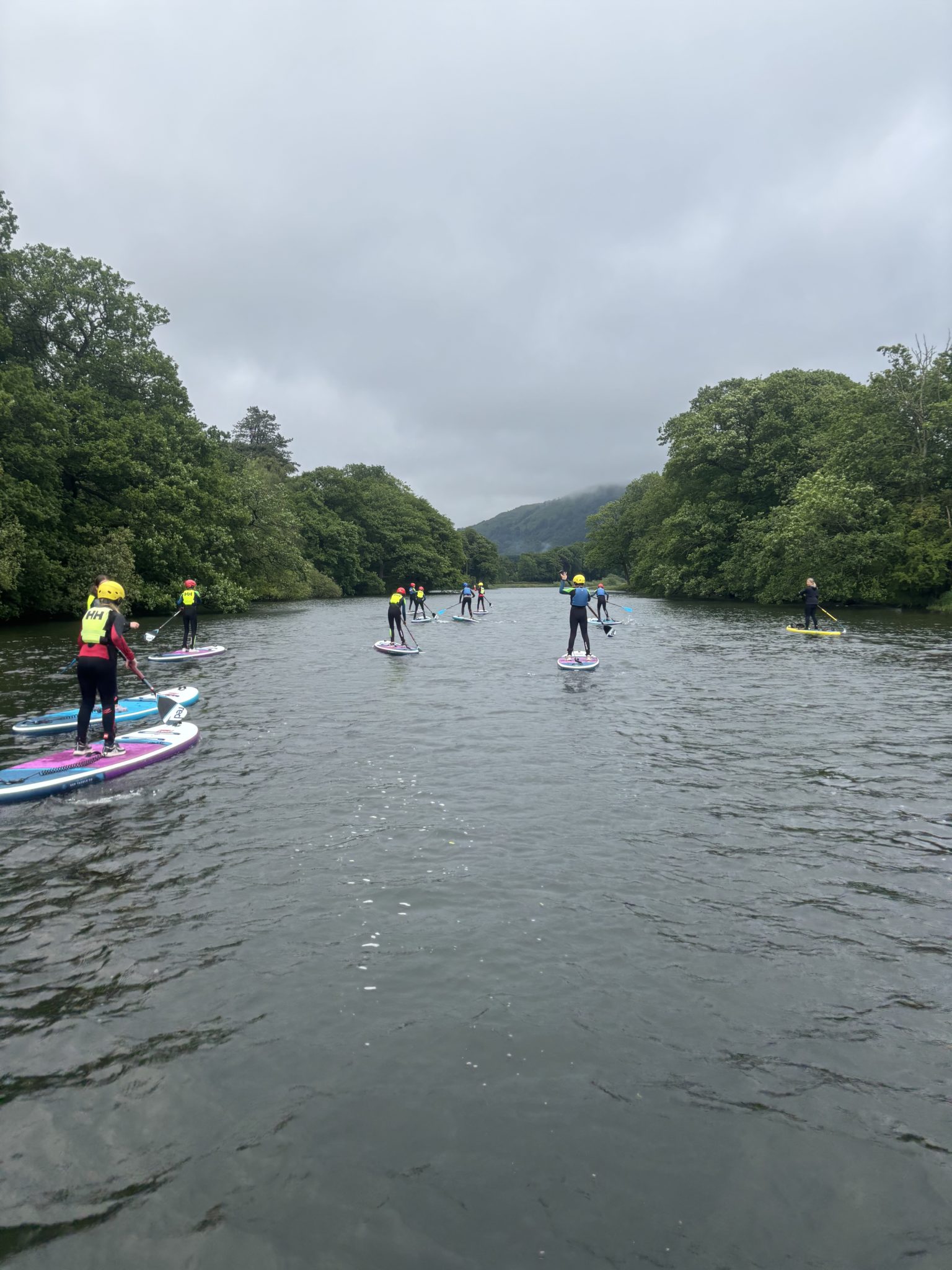 A group of people in wetsuits and helmets are paddleboarding on a wide, calm river surrounded by lush green trees under a cloudy sky.