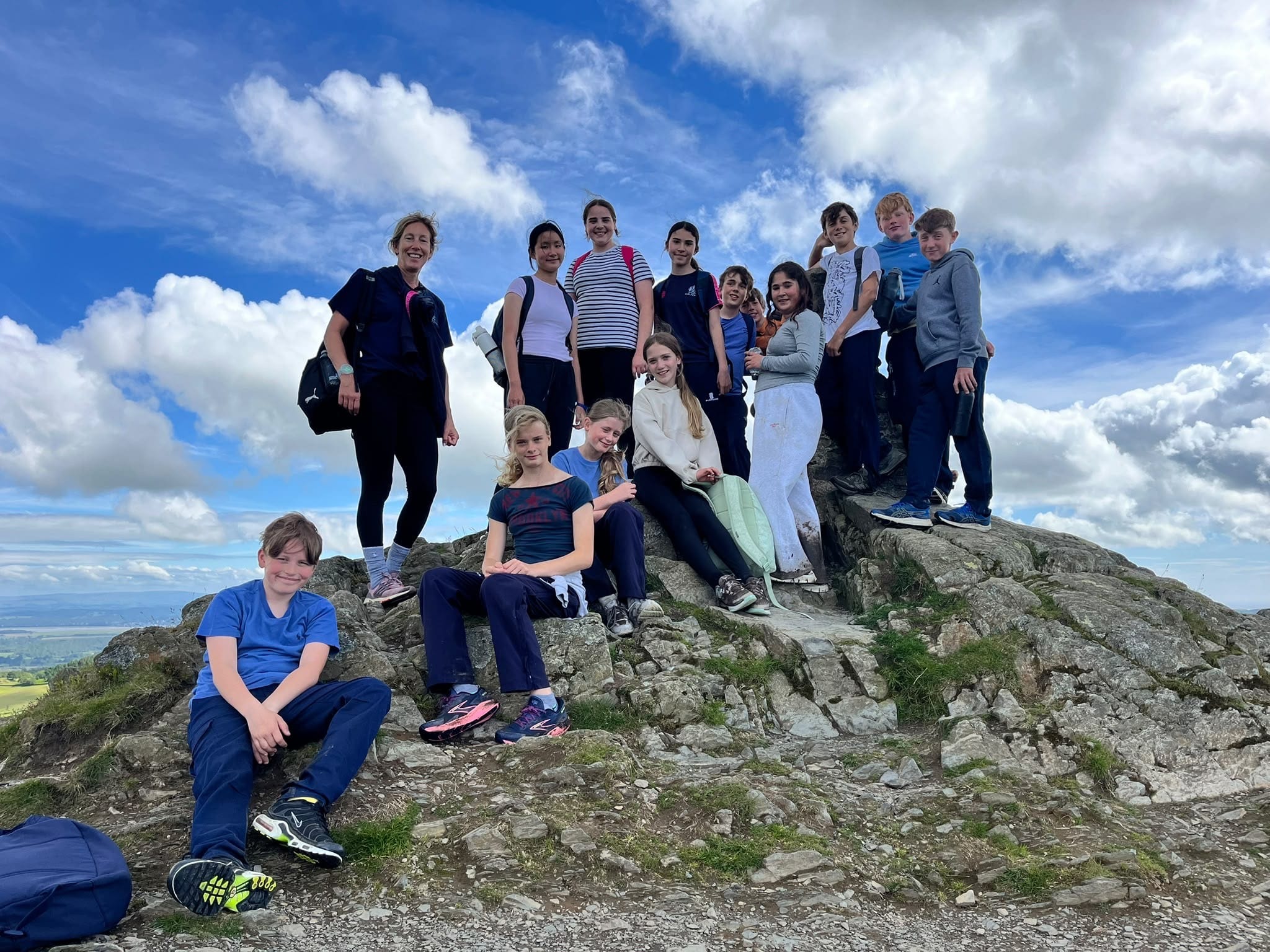 A group of people, mostly children, pose and smile on a rocky hilltop under a partly cloudy blue sky, with green fields visible in the distance.
