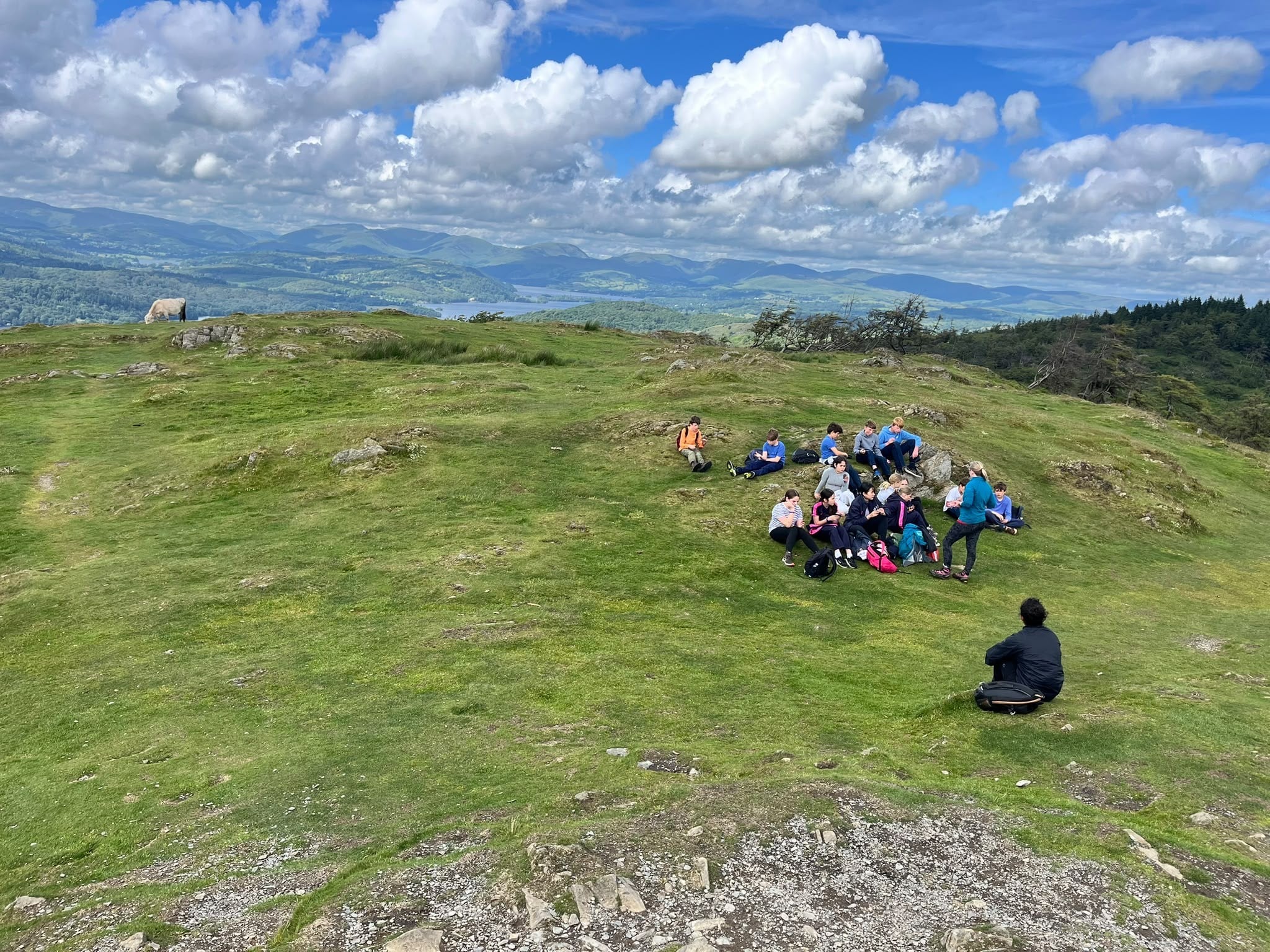 A group of people sit on a grassy hillside under a blue sky with scattered clouds, surrounded by rolling green hills and distant mountains. One person sits alone facing the group.