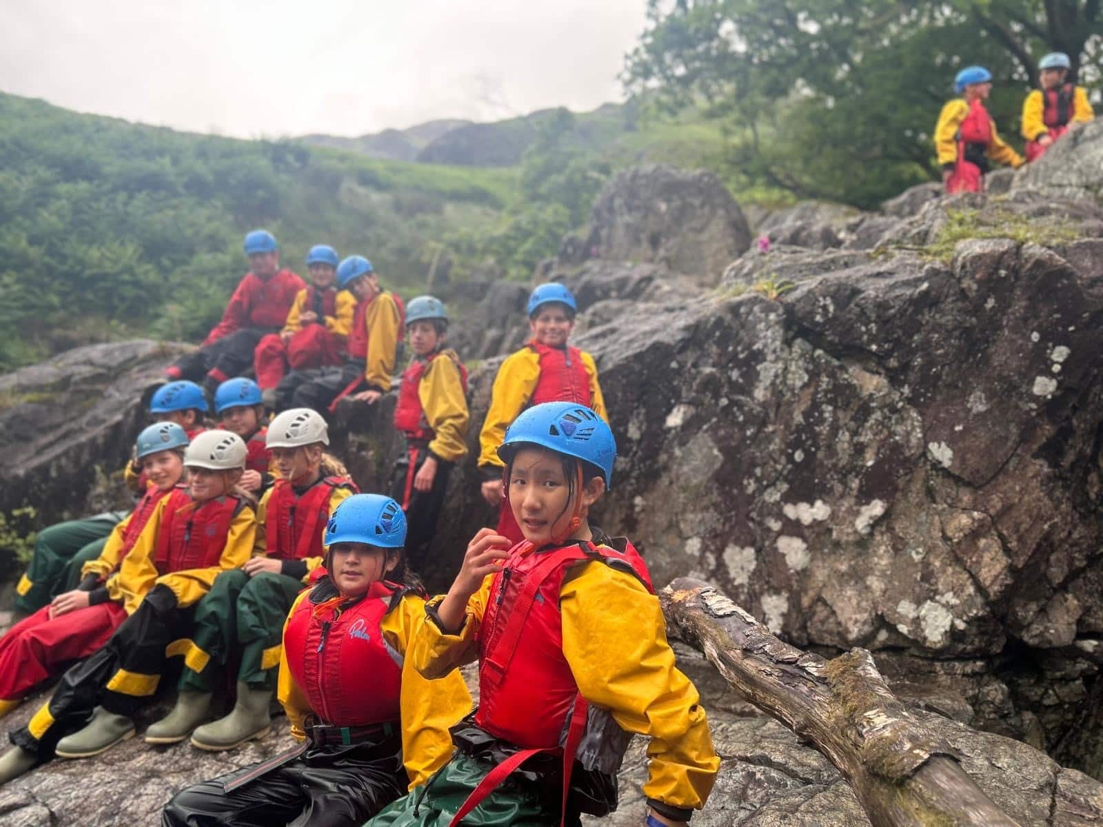 A group of children in colourful helmets and life jackets sit on rocks outdoors, surrounded by greenery. Some are smiling at the camera, suggesting they are on an adventure or outdoor activity.