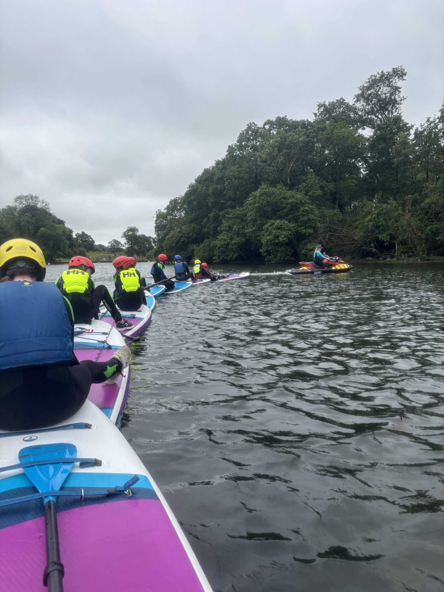 A group of people in colourful life jackets and helmets paddle on surfboards in a line along a river, with lush green trees on the banks under a cloudy sky.