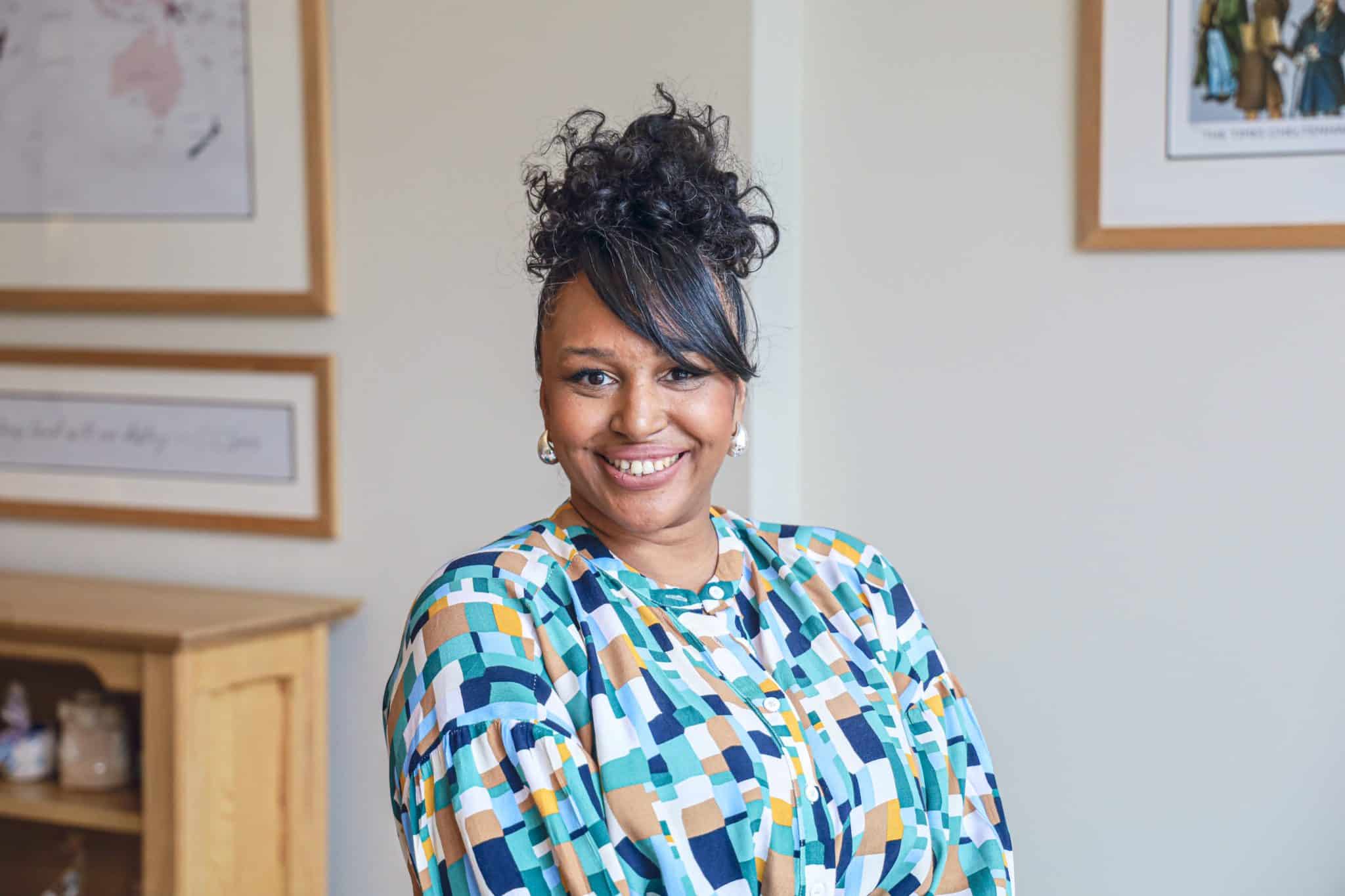 A woman with curly hair put up, wearing a colourful, patterned blouse, smiles at the camera in a well-lit room with framed artwork and a wooden cupboard—perfect for sharing her experience with Cheltenham College admissions.