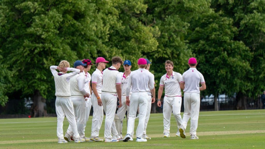 A group of young men in white cricket kit stand together on a grassy field, some wearing pink caps, appearing to celebrate or strategise, with trees in the background—capturing the spirited camaraderie that defines Cheltenham College, recently recognised in the Independent Schools of the Year Awards 2026.