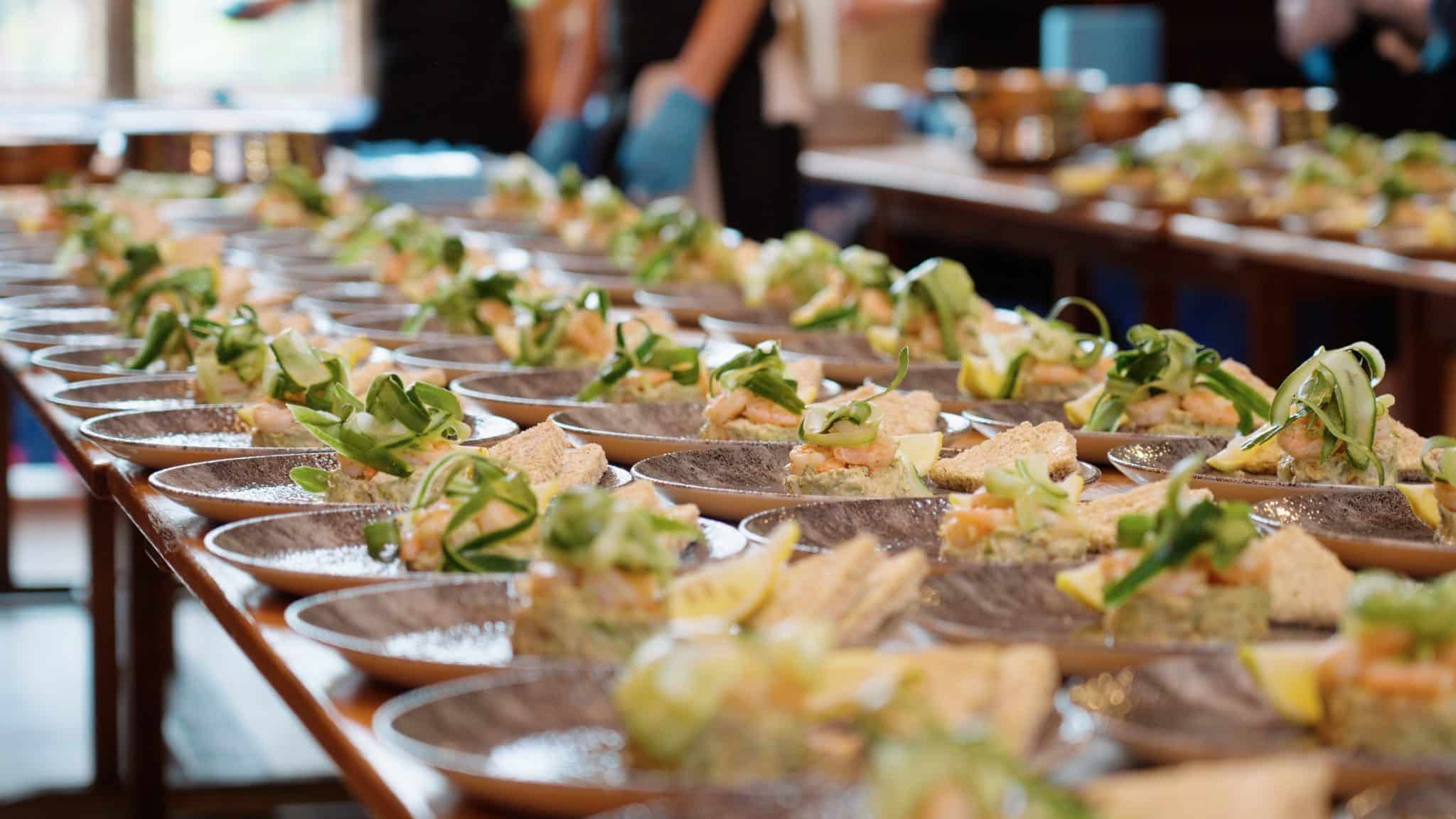 Rows of plates with neatly arranged appetisers topped with greens and lemon wedges sit on a long wooden table, ready to be served at an event; blurred figures are in the background.