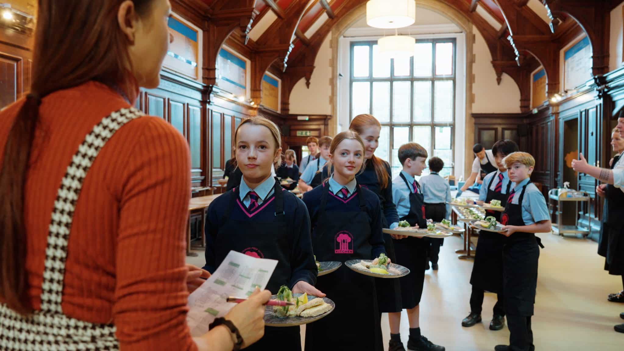 Children in school uniforms carry plates of food in a large, wood-panelled hall. An adult stands in the foreground holding a piece of paper, interacting with the children. Other adults and pupils are visible in the background.