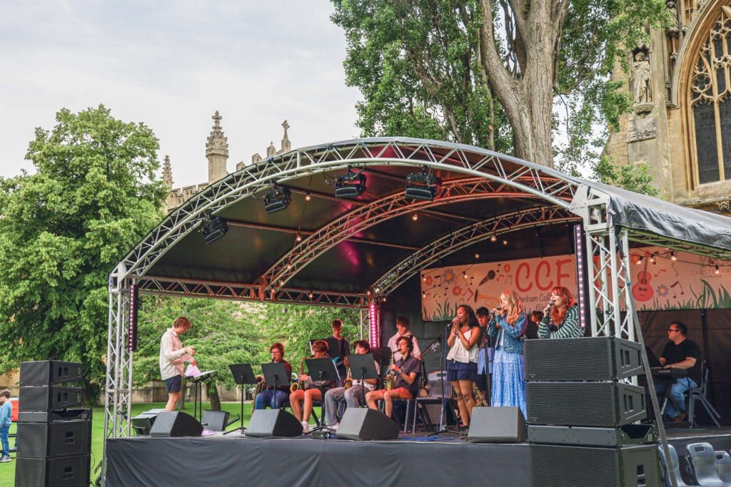 A band performs on an outdoor stage under a canopy at Cheltenham College, with musicians playing instruments and singers at microphones. Trees and a stone building are visible in the background.