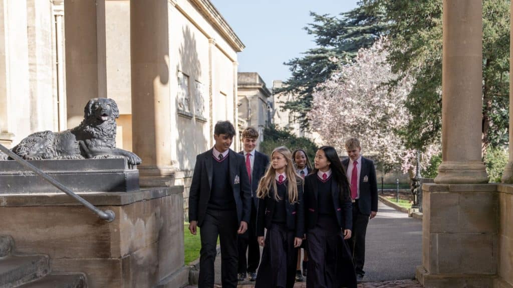 A group of students in uniforms walk together outside a historic building, passing a statue of a dog. Classical roots are evident in the stone columns and timeless architecture that frame their path, while these modern minds move purposefully beneath the trees.