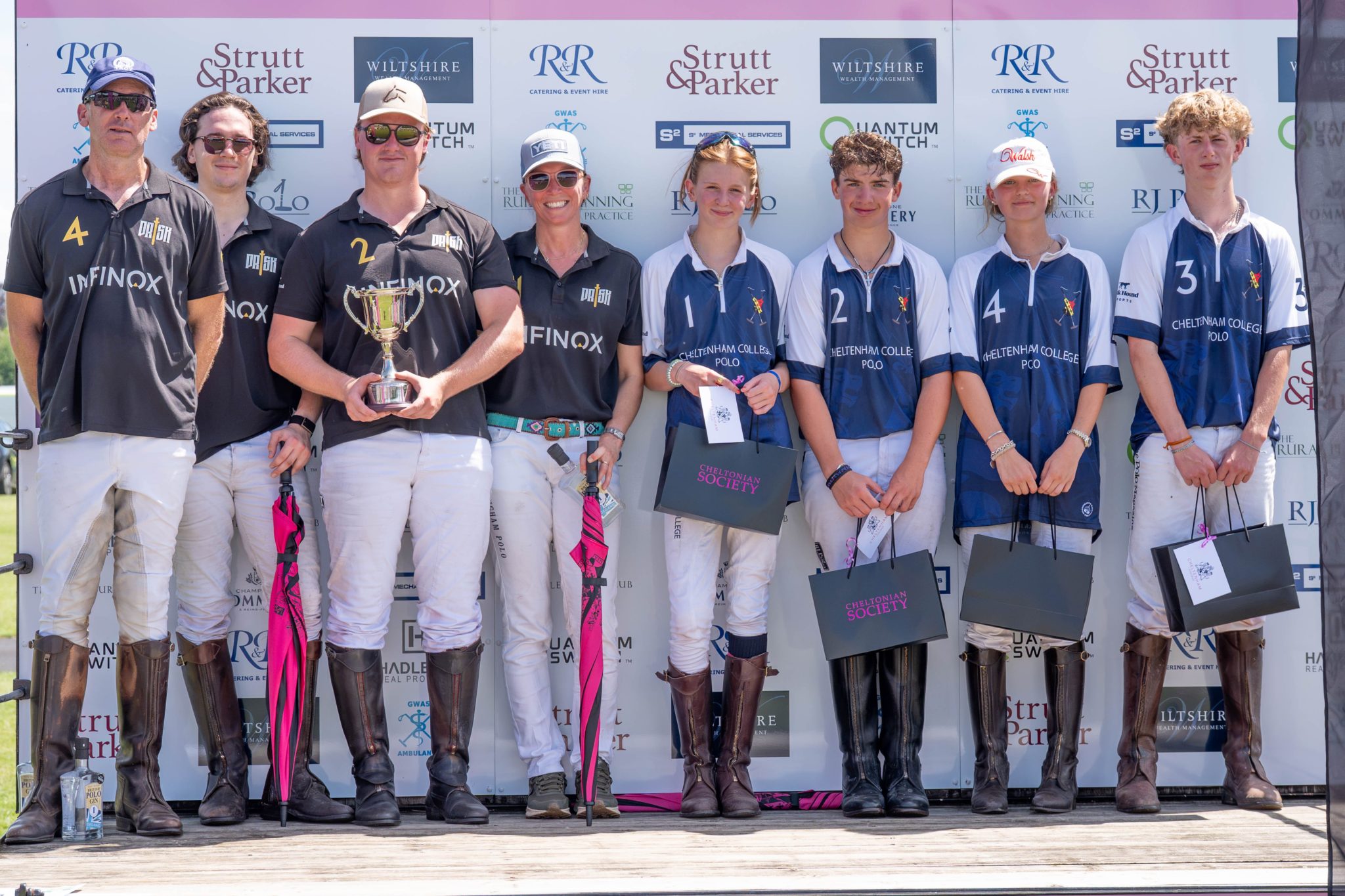 Eight young polo players in riding kit and boots stand on a podium holding trophies and gift bags, posing in front of a sponsor backdrop at an awards ceremony. Four wear black, four wear blue and white kits.