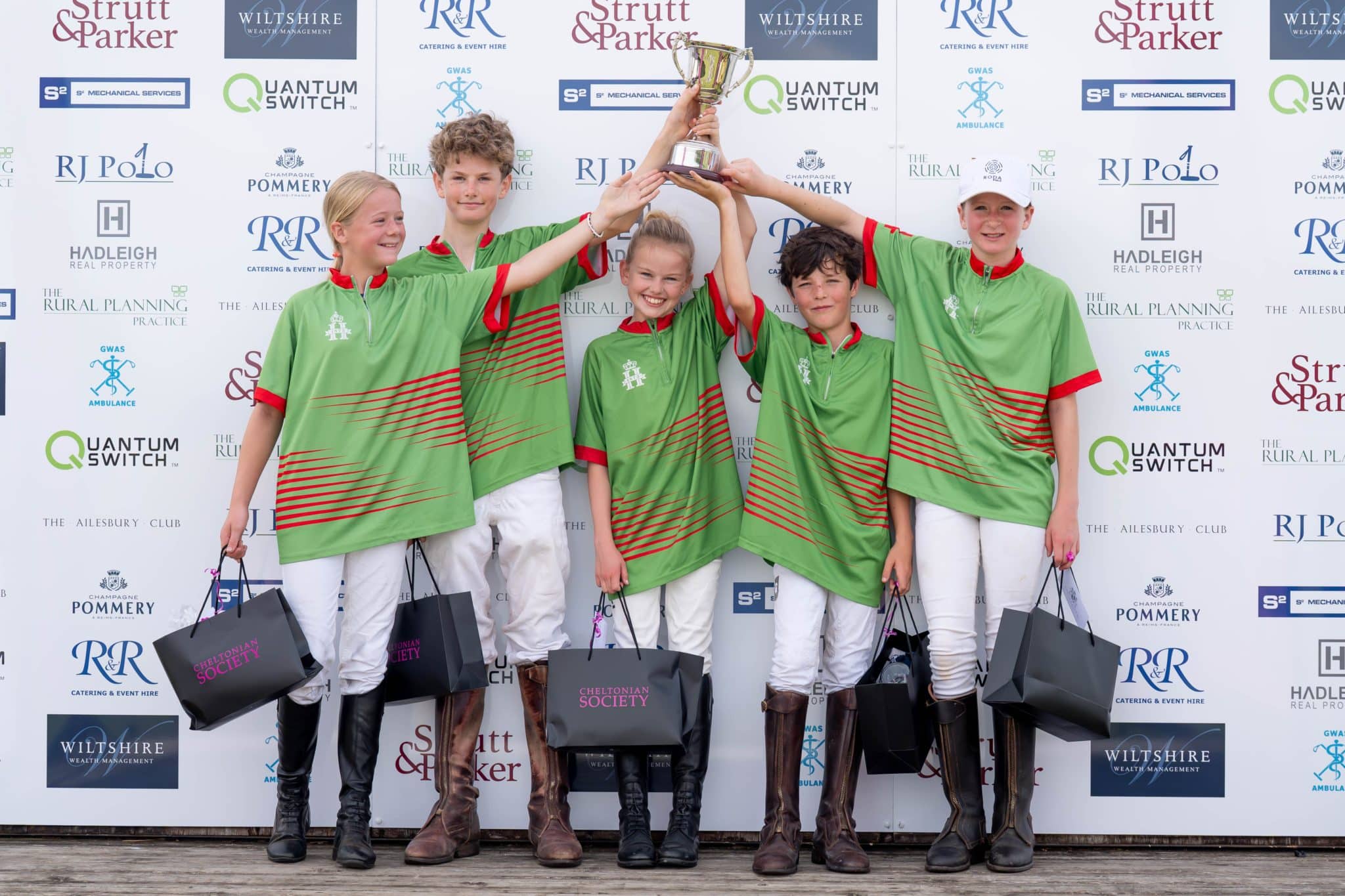 Five young polo players in matching green uniforms stand in front of a sponsor backdrop, smiling and holding a trophy above their heads. They each carry gift bags and wear riding boots and white trousers.