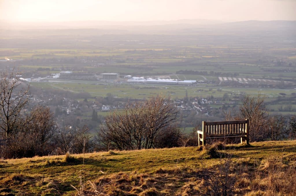 A wooden bench on a grassy hilltop overlooks a rural landscape with fields, trees, a village, and distant hills under a hazy sky, reminiscent of the tranquil views surrounding Cheltenham College.