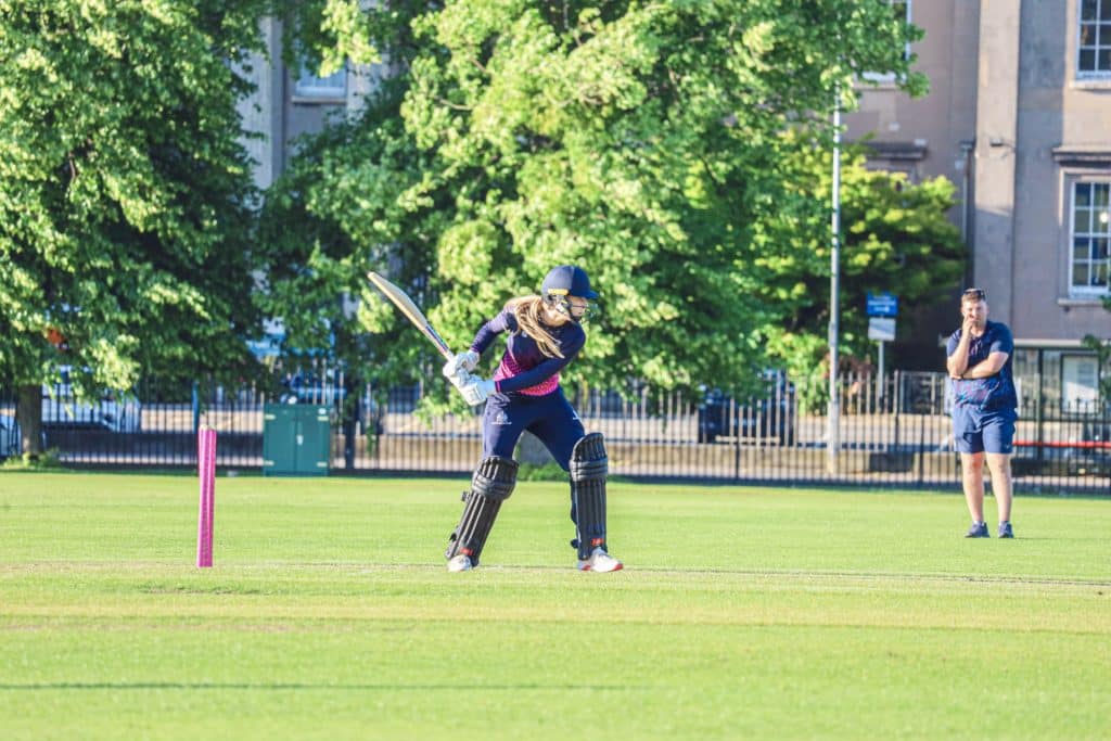 A cricket player in full kit prepares to hit the ball on a sunny outdoor pitch at Cheltenham College, whilst another person in casual clothes watches from a distance.