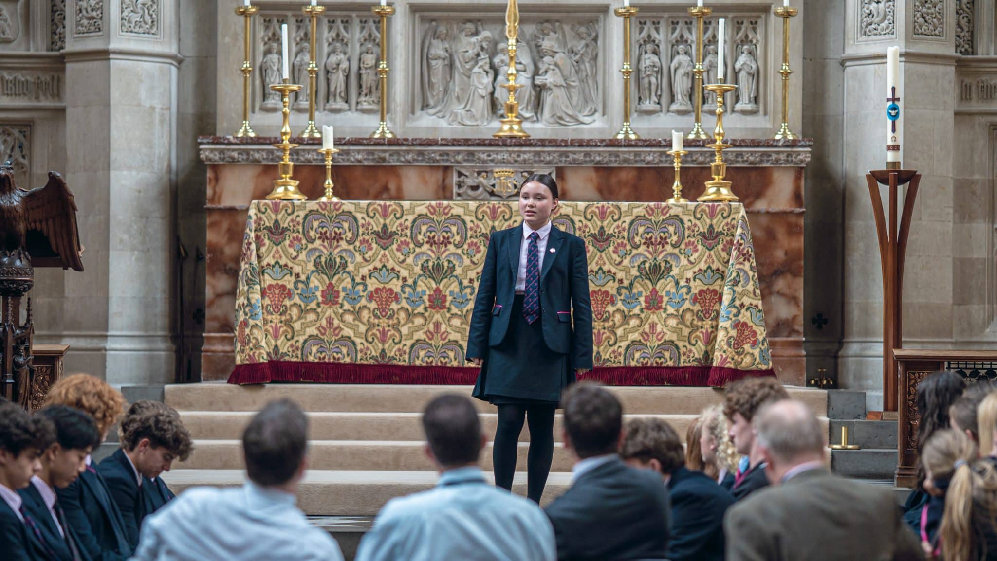 A student in a school uniform stands speaking in front of an ornate altar in a church or chapel, whilst a group of people seated below listen attentively.